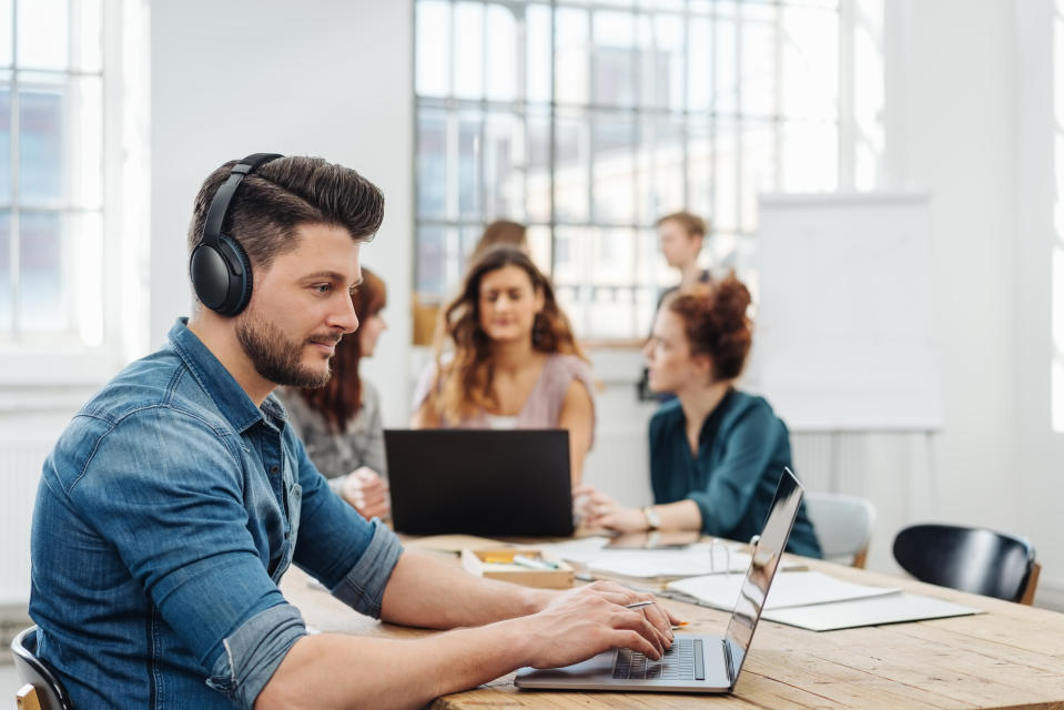 Modern office with a dedicated young business team working on paperwork and laptops seated around a table with focus to a young man wearing headphones typing on a laptop