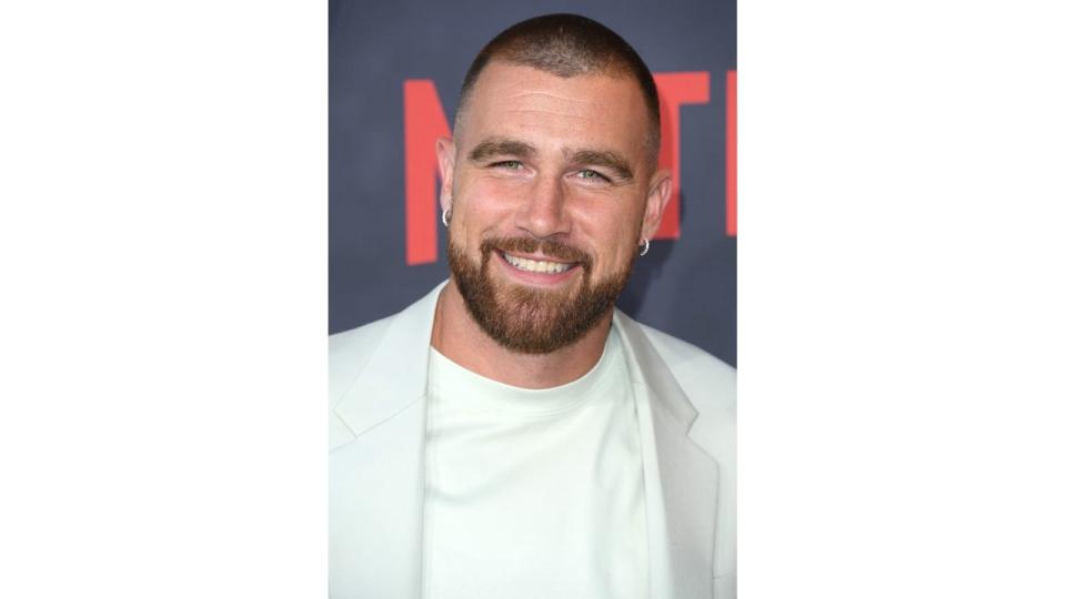 Travis Kelce smiling in a white suit jacket 