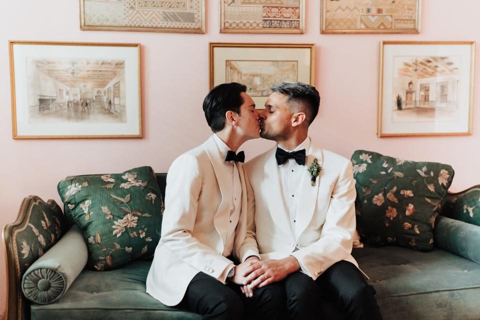 Two grooms in white tuxedo jackets kiss on a green couch.