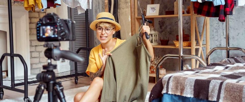 Young woman fashion blogger influencer wearing hat and eyeglasses sitting on fluffy carpet at urban stylish apartment recording video vlog on digital camera telling clothing tips