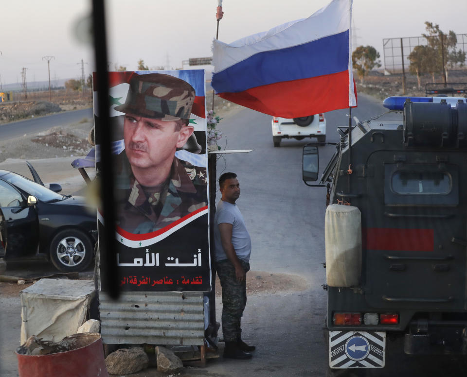 Syrian army soldier stands at a check-point as Russian military police vehicle, right, passes by near the village of Almajdiyeh, Syria, Tuesday, Aug. 14, 2018. The Russian military said Tuesday that its forces in Syria will help U.N. peacekeepers fully restore patrols along the frontier with the Israeli-occupied Golan Heights, reflecting Moscow's deepening role in mediating between the decades-old foes. (AP Photo/Sergei Grits)