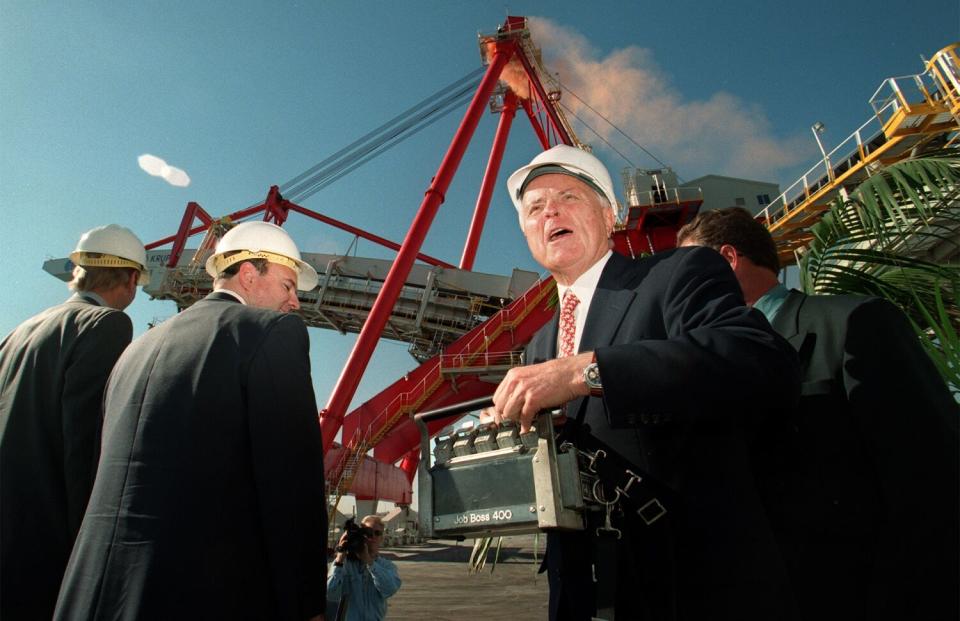 A man in a business suit and hard hat works the controls of a loading crane