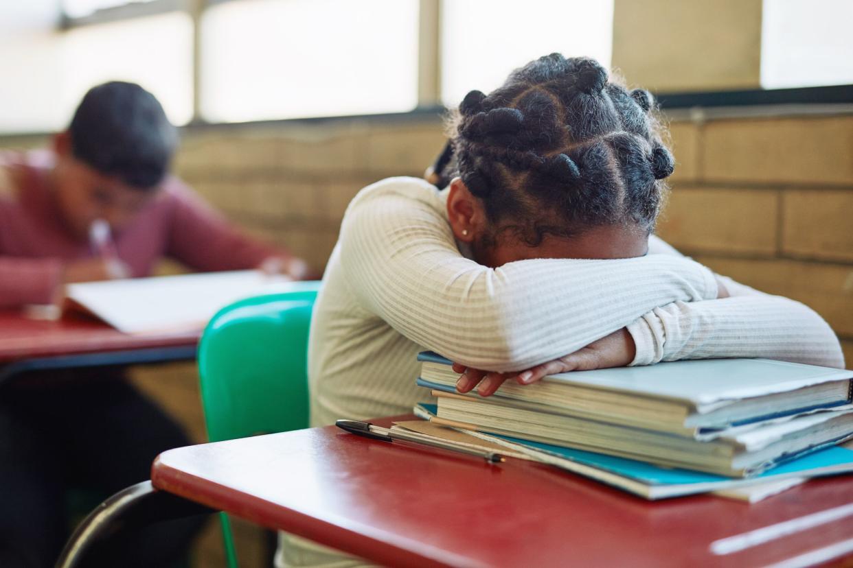 Young girl covering her head in her folded arms on a pile of school books on a red student desk with a green chair in elementary school, next to a brick wall with windows, and a young boy at his desk working blurred, behind her