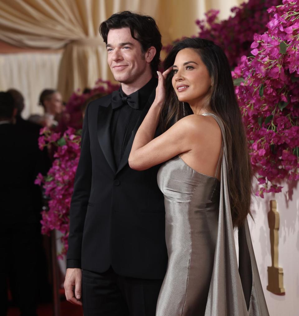 John Mulaney, in a black suit and tie, and Olivia Munn, in a silver gown, pose for cameras.