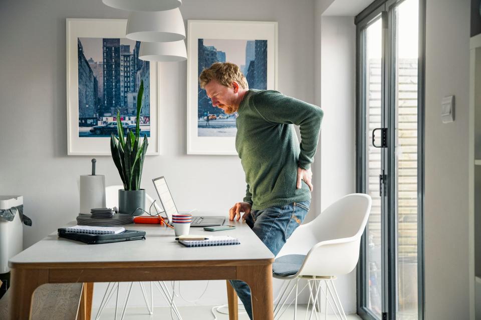 A man stands up from his desk and clutches his lower back in pain