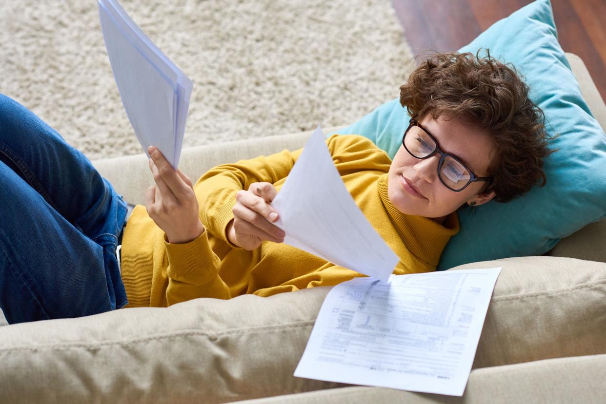 serious concentrated beautiful young woman in glasses lying on sofa and analyzing papers while preparing tax return at home