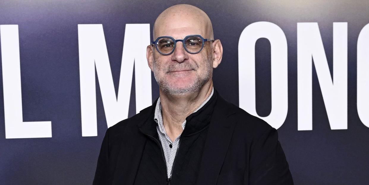 harlan coben, a man stands looking at the camera smiling, he wears glasses, a white shirt and black jacket