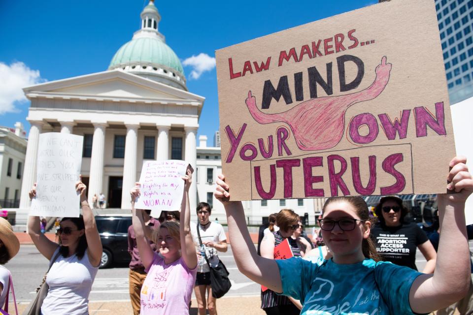 Demonstrators standing in front of a government building hold signs that read "lawmakers, mind your own uterus"