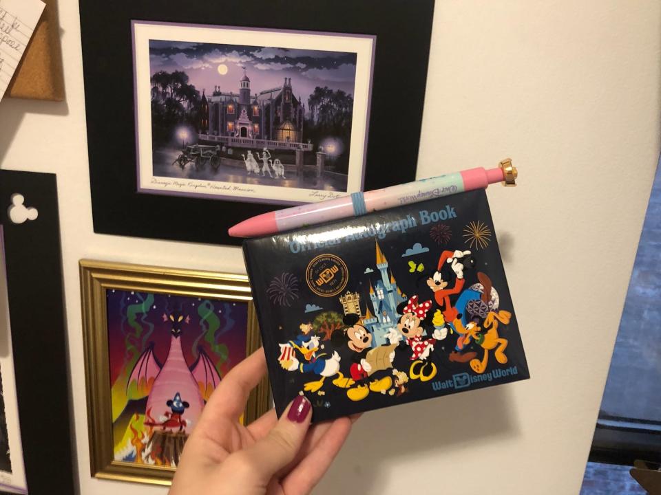 hand holding an official disney autograph book with a novelty pen in front of some disney art