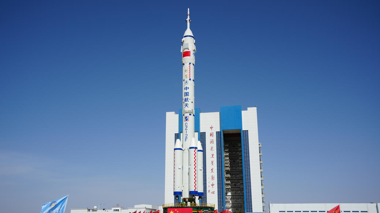  A tall white rocket with a red square near the top and chinese writing on the side, stands outside a hanger building beneath a blue sky. 