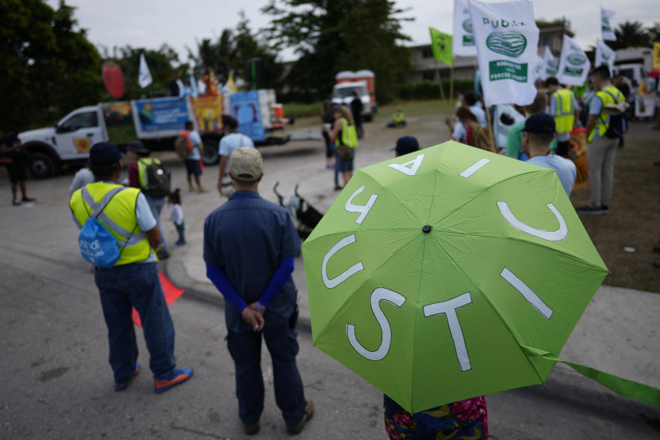 A woman holds an umbrella reading "Justice," in Spanish, as farmworkers and allies prepare to start a five-day trek aimed at highlighting the Fair Food Program, which has enlisted food retailers to use their clout with growers to ensure better working conditions and wages for farmworkers, Tuesday, March 14, 2023, in Pahokee, Fla. (AP Photo/Rebecca Blackwell)