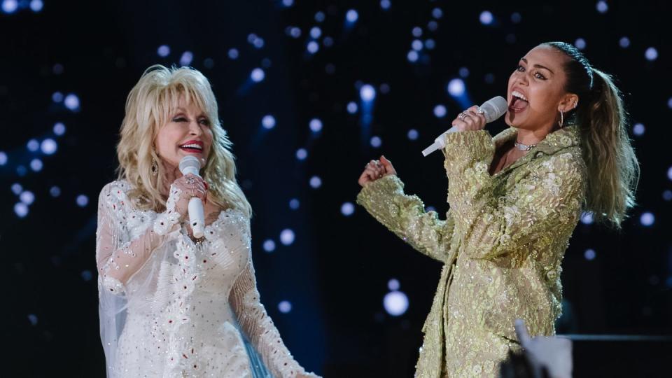 dolly parton, wearing a white dress, and miley cyrus, wearing a gold outfit, as both sing into handheld microphones on a stage, with blue lights behind them