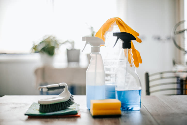 Cleaning supplies including spray bottles with cleaning liquid, scrub brush, sponge, cleaning towels, and yellow rubber gloves on a table