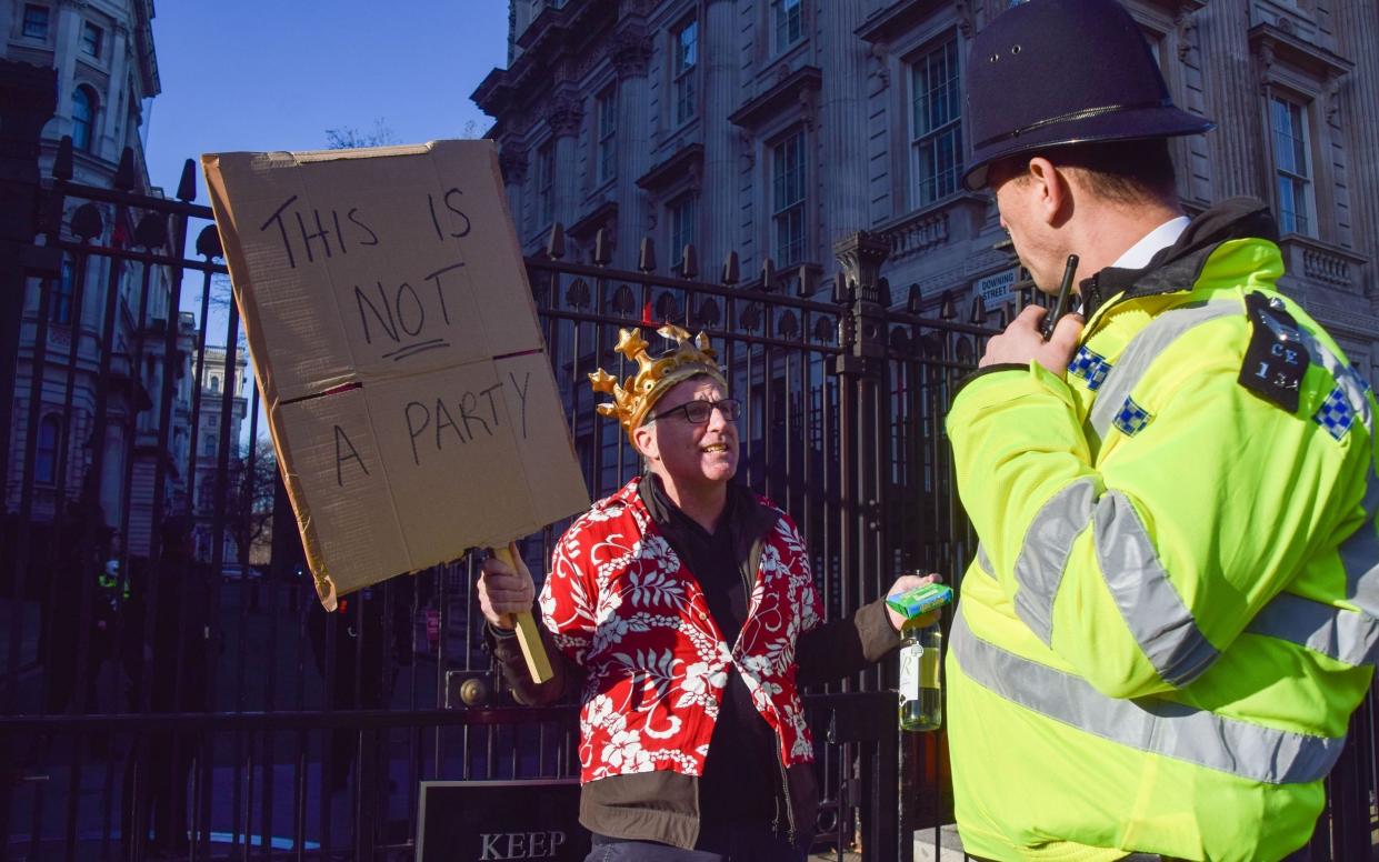 A protester wearing a crown and holding a packet of cheese, a bottle of wine and a 'This Is Not A Party' placard speaks to a police officer during the demonstration outside Downing Street - Vuk Valcic 