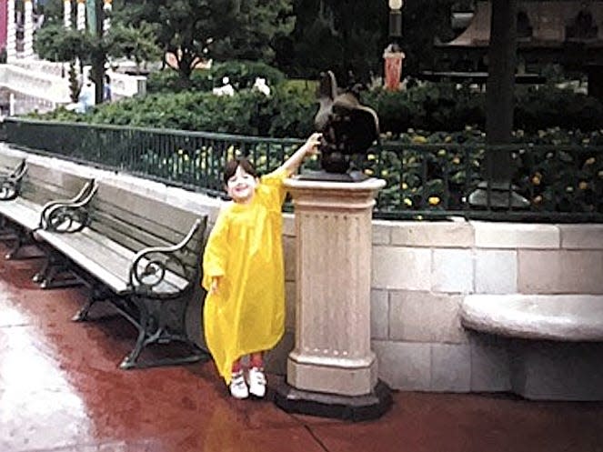 megan as a kid at disney world 30 years ago wearing a yellow poncho and touching a dumbo statue