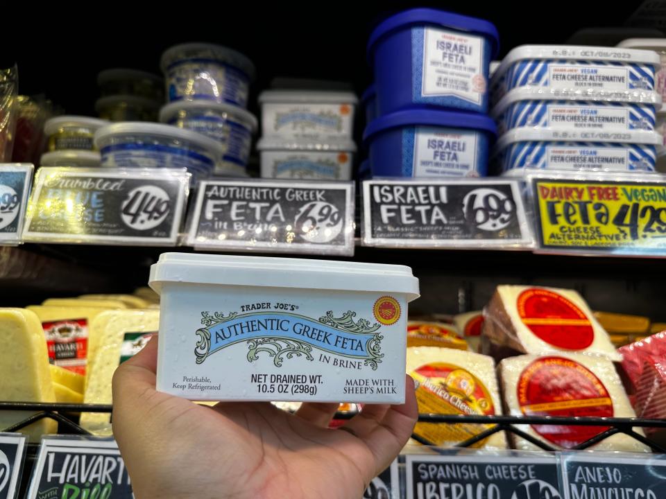 hand holding tub of feta cheese in front of a cheese fridge at trader joe's