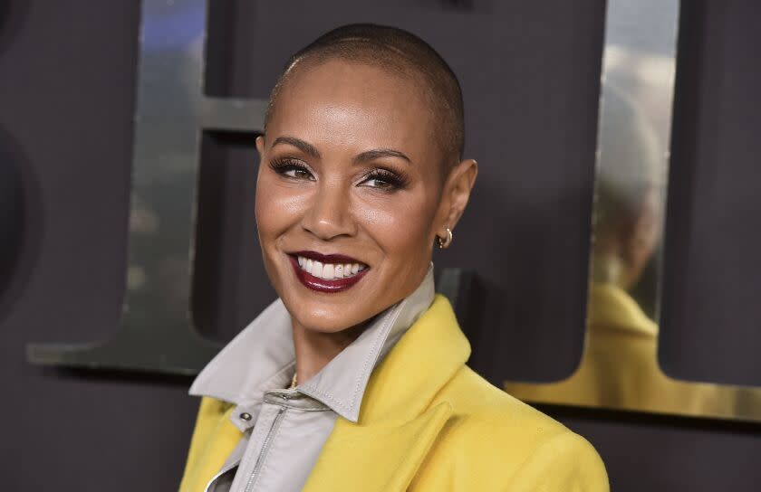 A woman with a shaved head smiling in dark red lipstick and a yellow jacket