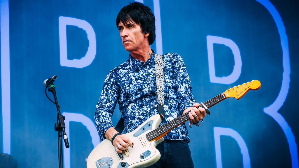 Johnny Marr, with short black hair and wearing a blue, white and black speckled shirt and blue jeans, plays a white guitar next to a microphone