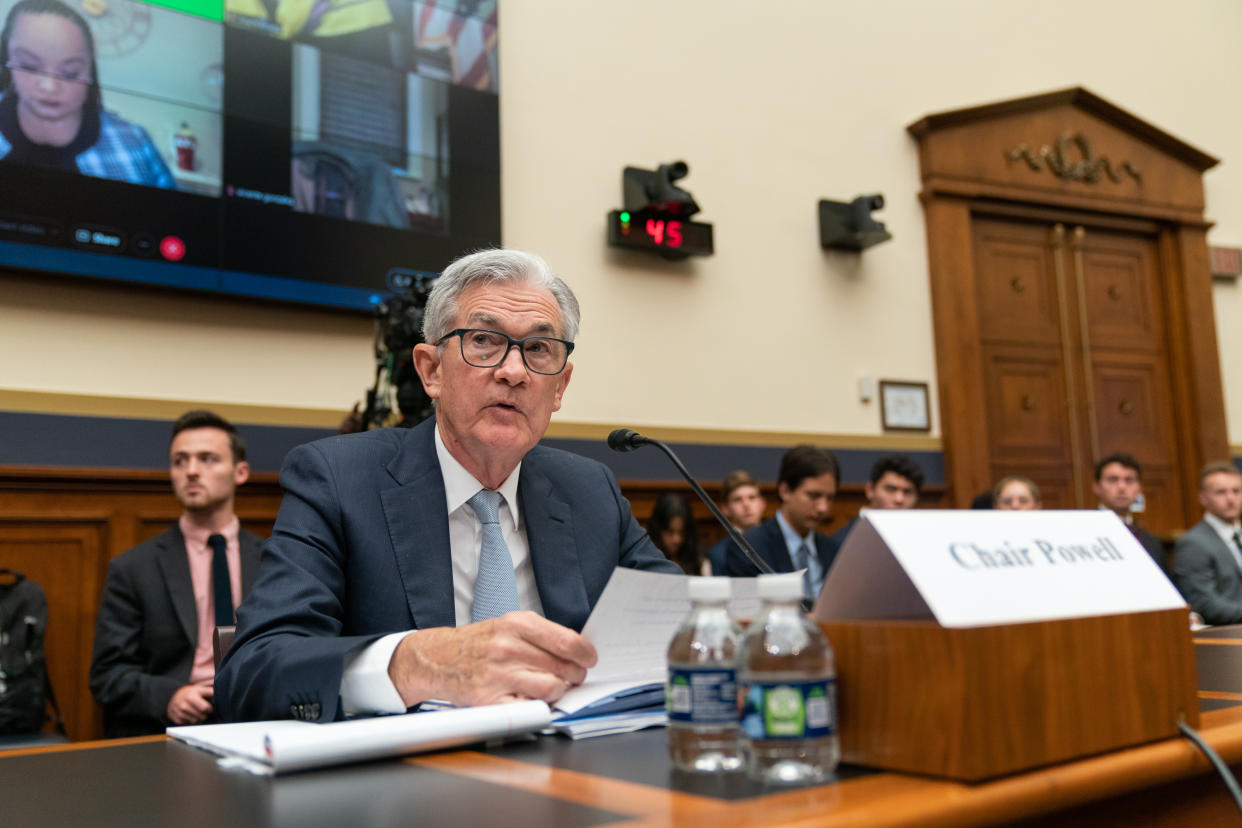Jerome Powell at a desk in a hearing, with a TV screen in the background.