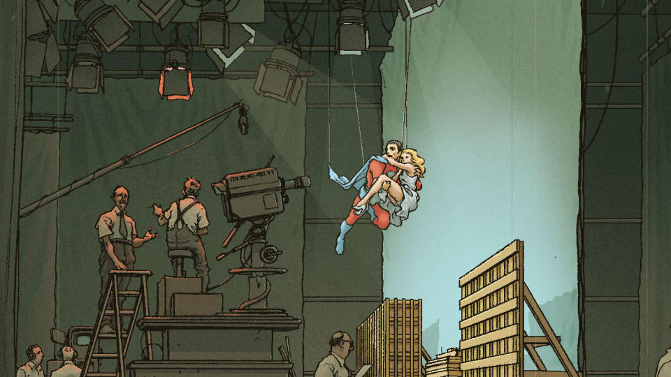 A film set. A superhero is winched down from the ceiling carrying a woman in his arms.