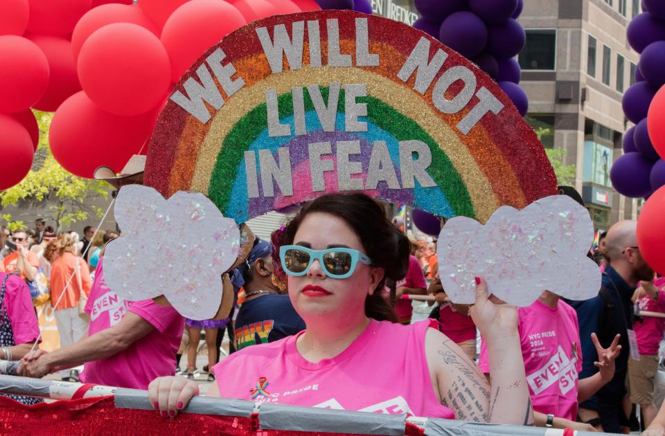 A protester in pink shirt and blue shades stands under an elaborate, rainbow Gay Pride banner with the text 