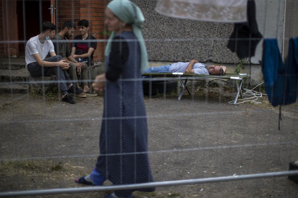 A woman walks past migrants from Iraq resting at the refugee camp in the village of Verebiejai, some 145km (99,1 miles) south from Vilnius, Lithuania, Sunday, July 11, 2021. Migrants at the school in the village of Verebiejai, about 140 kilometers (87 miles) from Vilnius, haven't been allowed to leave the premises and are under close police surveillance. Some have tested positive for COVID-19 and have been isolated in the building. (AP Photo/Mindaugas Kulbis)