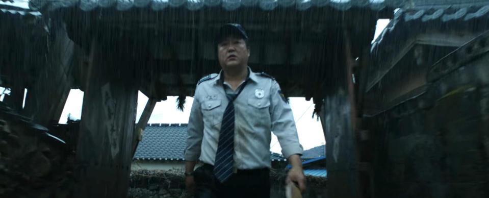 A man in a police uniform and tie walks beneath a traditional gate in the rain, holding an object in his hand