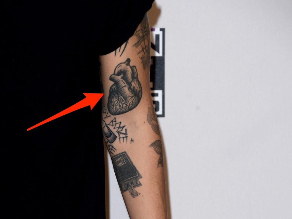 A red arrow pointing to an anatomical heart tattoo on Harry Styles' left arm.