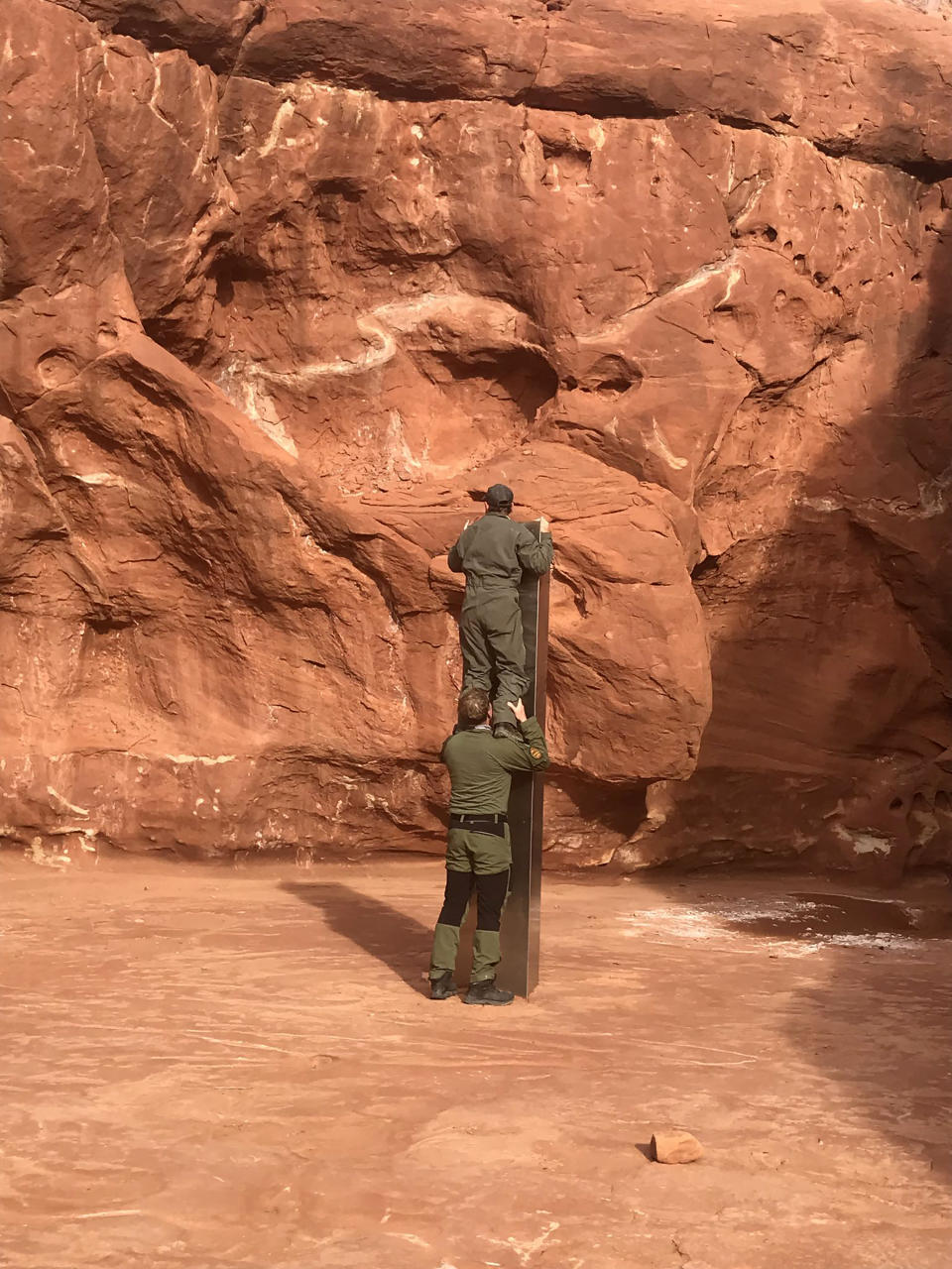 This Nov. 18, 2020 photo provided by the Utah Department of Public Safety shows Utah state workers checking out a metal monolith that was found installed in the ground in a remote area of red rock in Utah. The smooth, tall structure was found during a helicopter survey of bighorn sheep in southeastern Utah, officials said Monday. State workers from the Utah Department of Public Safety and Division of Wildlife Resources spotted the gleaming object from the air and landed nearby to check it out. The exact location is so remote that officials are not revealing it publicly, worried that people might get lost or stranded trying to find it and need to be rescued. (Utah Department of Public Safety via AP)