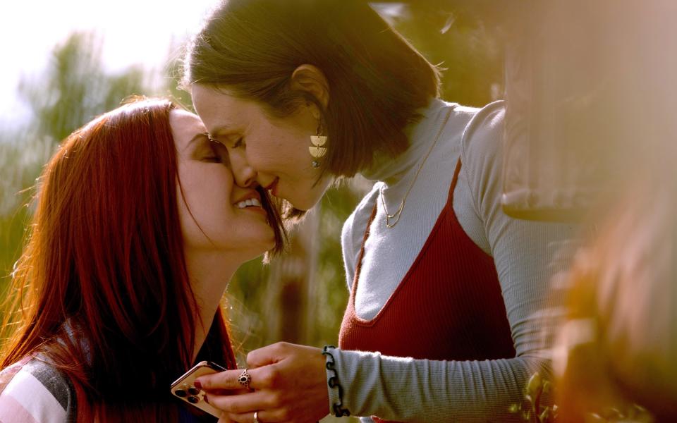 Two women share an intimate moment, smiling with their eyes closed as they lean in for a kiss. One holds a phone; the other wears heart-shaped earrings