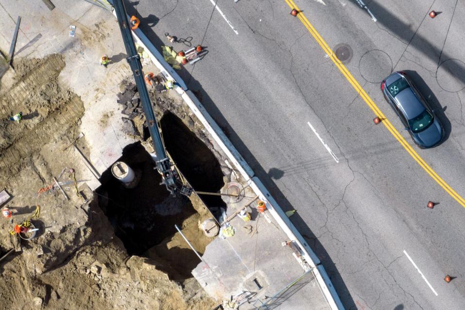 Crews work to repair a large sinkhole next to a four-lane road. A car drives by.