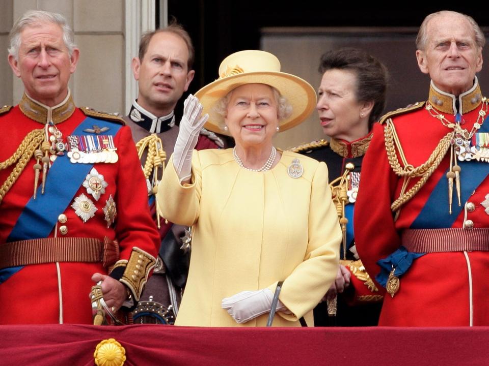 The Queen smiling and waving in a pale yellow overcoat with an oval silver brooch on it. She has a matching yellow wide-brimmed hat on, a necklace with pearls, and white gloves on.