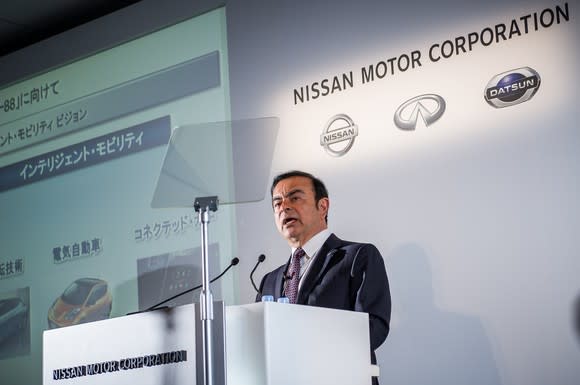 Ghosn is shown standing at a podium. Behind him, a backdrop with Nissan's logo.