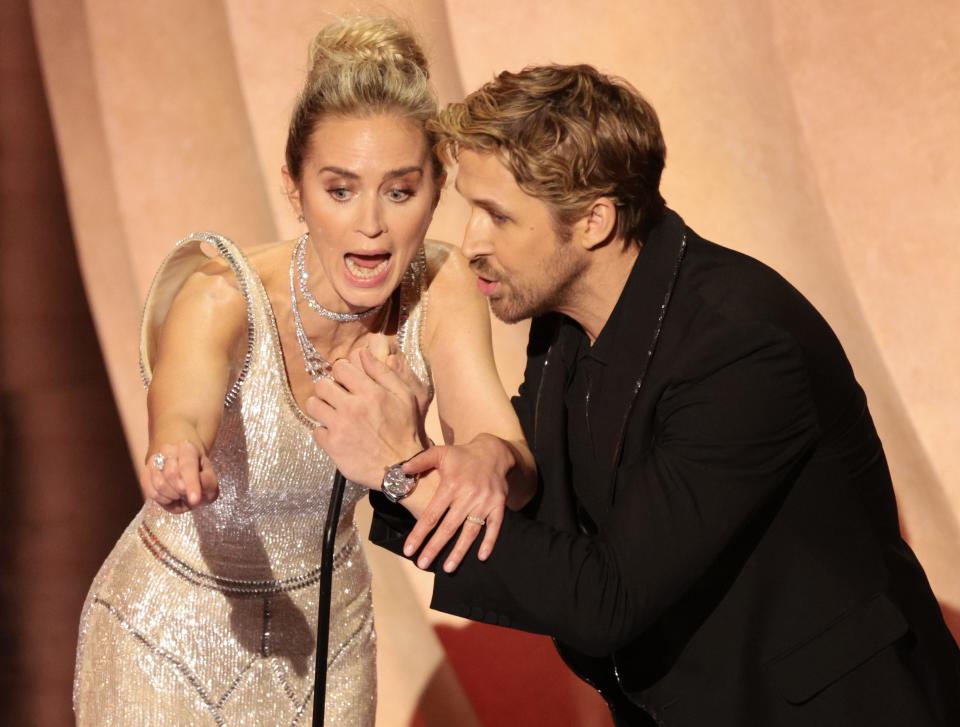 Emily Blunt pointing toward the audience and Ryan Gosling grabbing the microphone as they present at the Oscars.