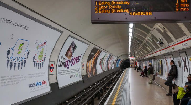 An image of indoor train tracks and platform with a curved wall, advertisements on the wall, people waiting on the platform, and a train schedule sign with orange text.