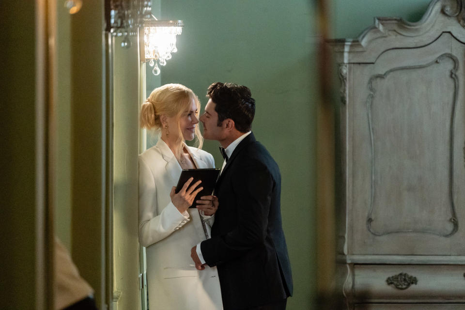 Jodie Foster and Shazad Latif are elegantly dressed, sharing an intimate moment in a refined room. Foster holds a small book, and Latif leans in to kiss her