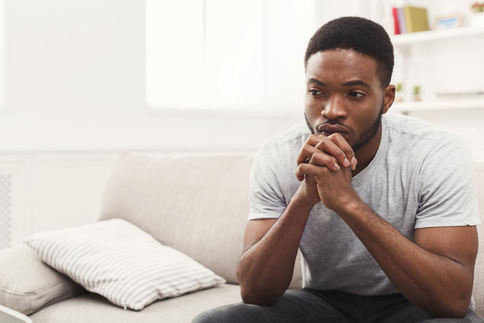 Man sitting on a sofa, leaning forward with his hands clasped, looking contemplative. No visible text or notable clothing style