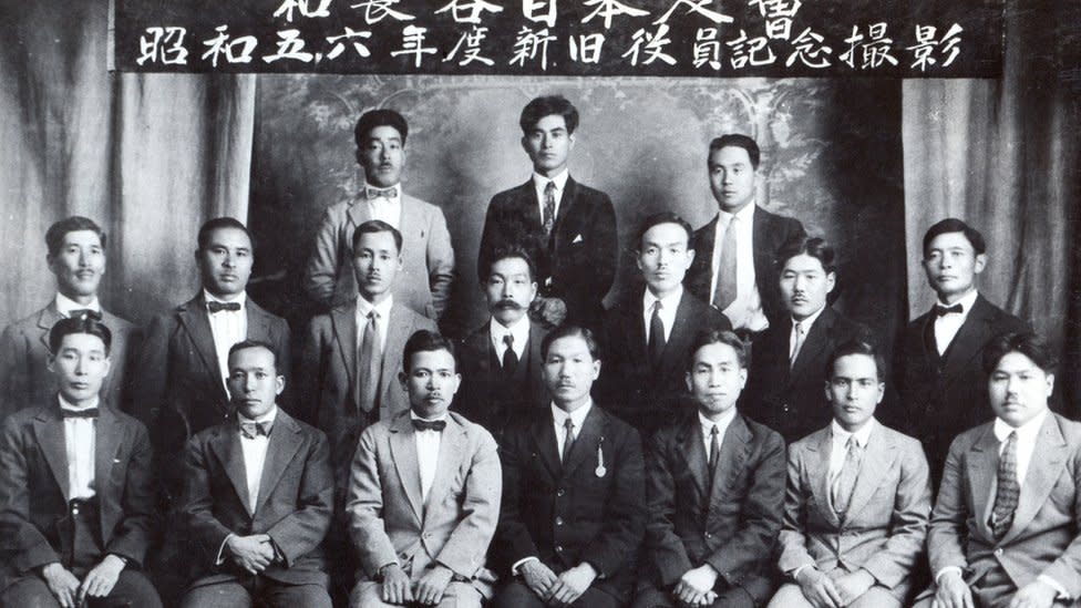 Hombres inmigrantes japoneses posan para una foto bajo un letrero en su idioma.