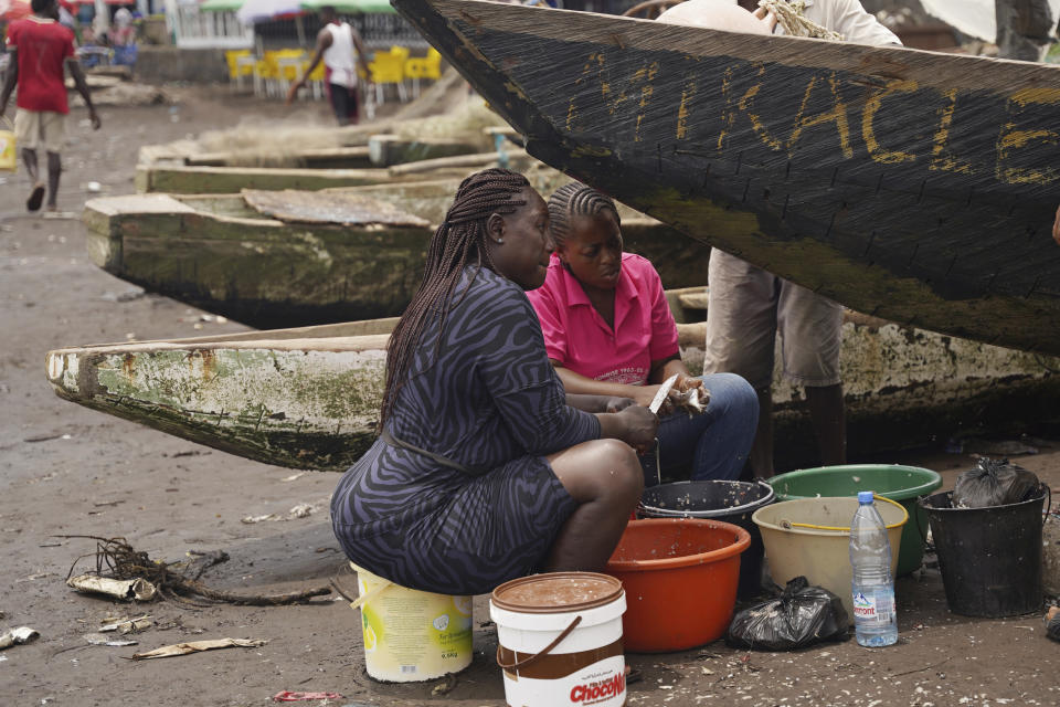 Fishmongers sit next to boats as they prepare freshly caught fish to be smoked on Limbe beach, Cameroon, on April 12, 2022. In recent years, Cameroon has emerged as one of several go-to countries for the widely criticized “flags of convenience” system, under which foreign companies can register their ships even though there is no link between the vessel and the nation whose flag it flies. But experts say weak oversight and enforcement of fishing fleets undermines global attempts to sustainably manage fisheries and threatens the livelihoods of millions of people in regions like West Africa. (AP Photo/Grace Ekpu)