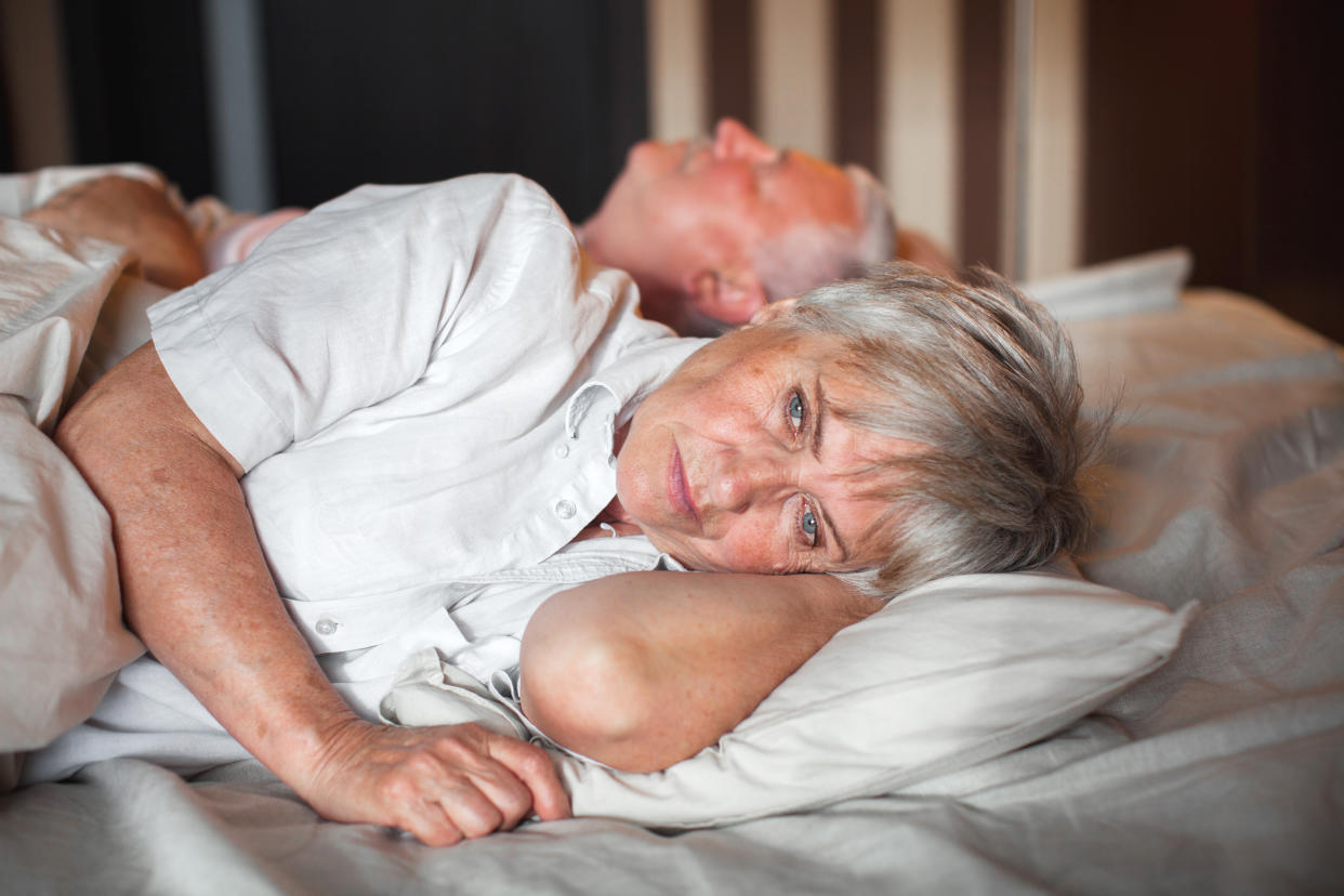 Upset mature woman lying in bed near her sleeping husband at home. Relationship problems
