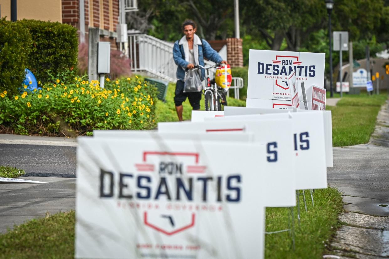 A person walks a bike on a sidewalk past campaign signs supporting Florida Governor Ron DeSantis.