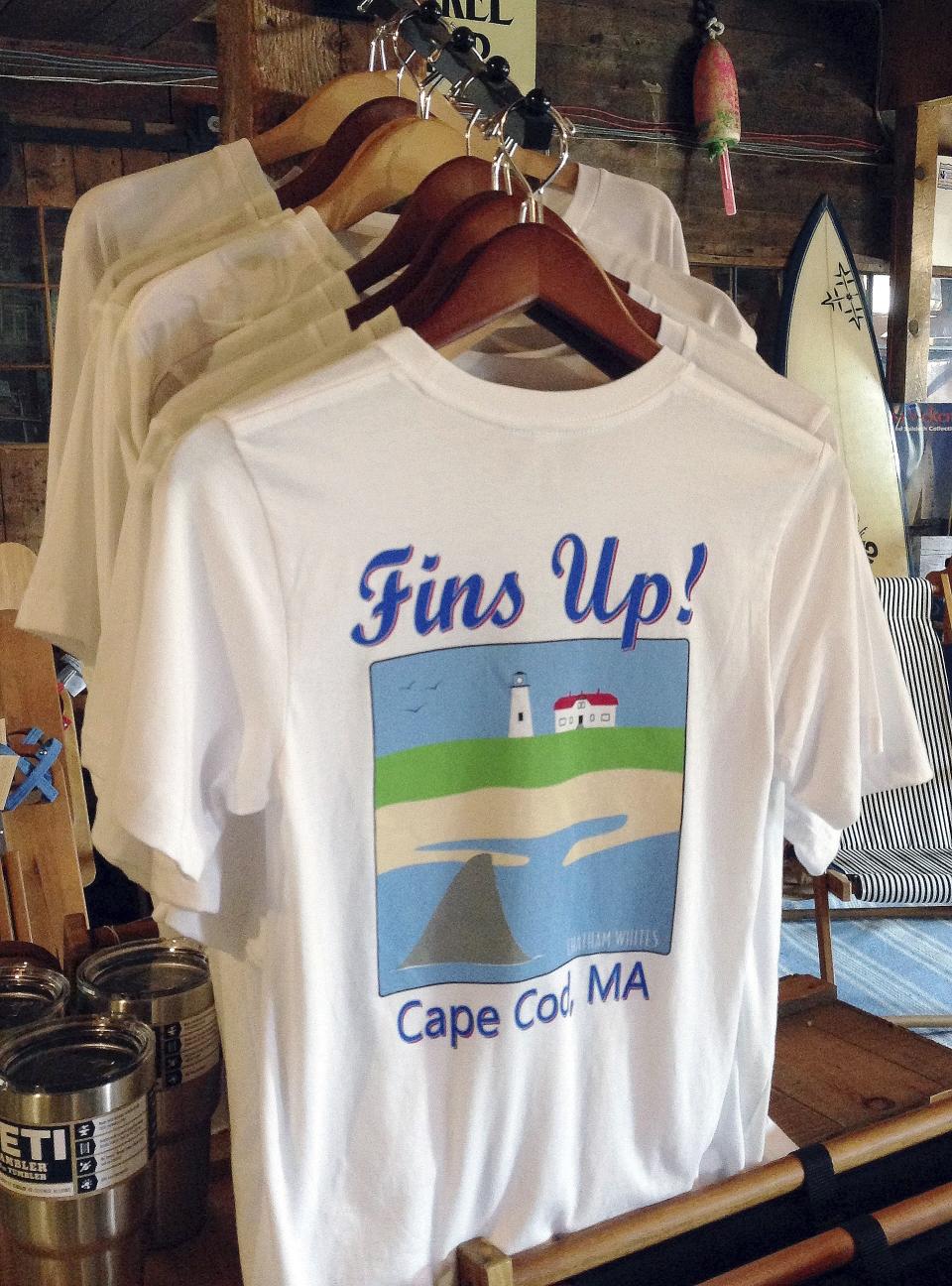 FILE - In this May 25, 2016 file photo, a shark-themed T-shirt is on sale at a souvenir shop in Harwich, Mass. Local chamber of commerce data suggests Cape Cod lodging and beach visit numbers are down in summer 2019, after uncommon tornados hit just one year after a pair of shark attacks. (AP Photo/Philip Marcelo, File)