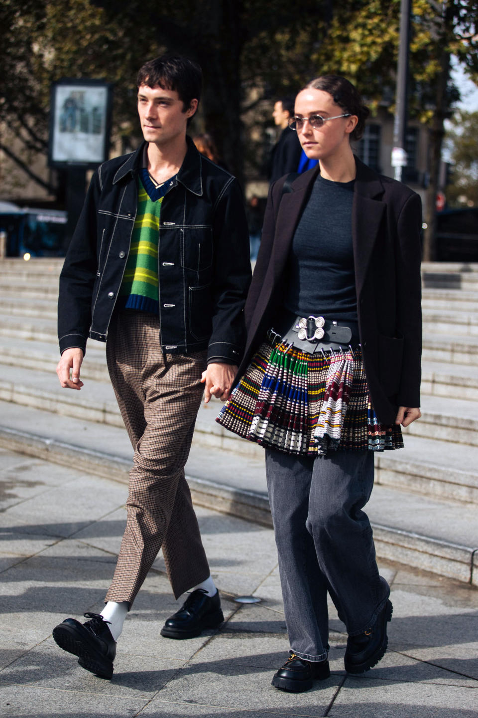 PARIS, FRANCE - OCTOBER 03: Editor Samuel Hine and Artist/Model Ella Emhoff attends the Ludovic de Saint Sernin show on October 03, 2021 in Paris, France. Sam wears a denim blue jacket, green striped cardigan top, brown gingham pants, white socks, and black oxford loafers. Ella wears a purple blazer, black top, Chopova Lowena colorful midi beaded pleated skirt with a black belt, black pants, and black boots. (Photo by Melodie Jeng/Getty Images)