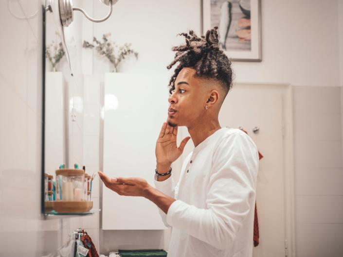 A young man in profile looks in the mirror as he applies skincare product to his face.