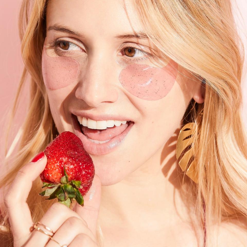 Model with pink eye gel patches applied to fact, smiling and holding a strawberry