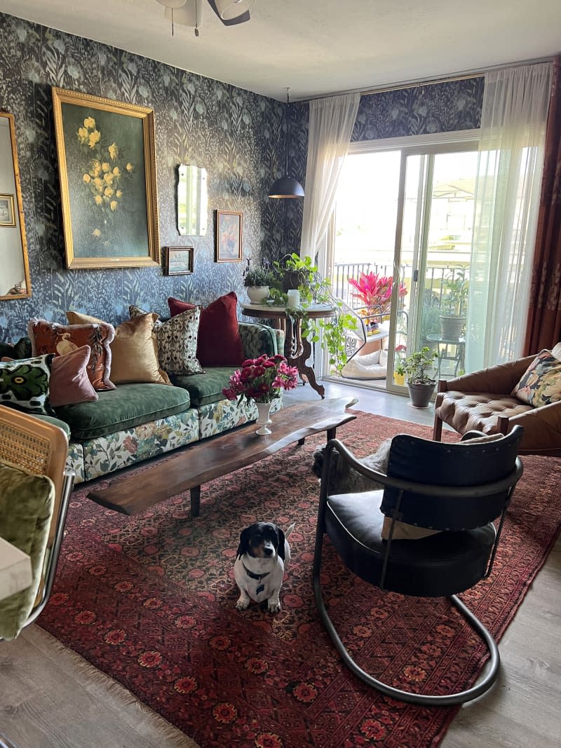 Living room with botanical wallpaper, green floral sofa and pillows, and lots of patterned textiles. Dog on the rug