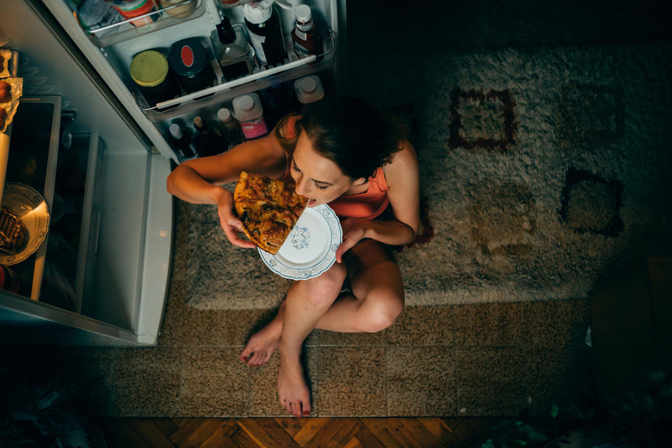 A person is sitting on the floor in front of an open refrigerator, eating pizza from a plate