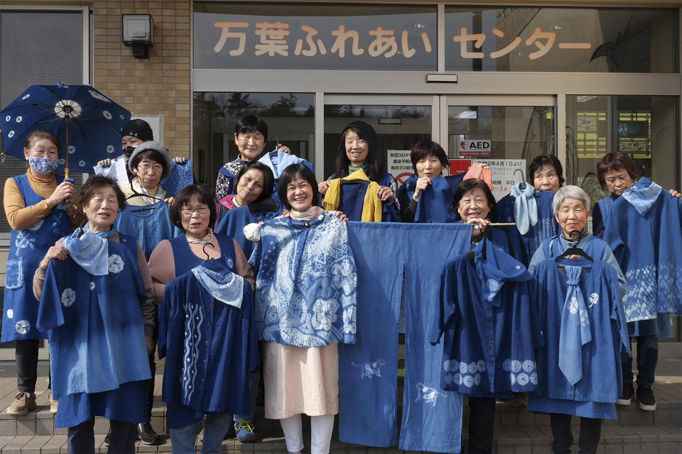 In this image from video, members of Japan Blue hold up their dyed clothes and pose for a photo in front of a community center which was used as an evacuation center when the massive earthquake hit the area in 2011, in Minamisoma, Fukushima Prefecture, northeastern Japan, on Feb. 20, 2021. Because of radiation released by the Fukushima nuclear plant disaster a decade ago, farmers in nearby Minamisoma weren't allowed to grow crops for two years. After the restriction was lifted, two farmers found an unusual way to rebuild their lives and help their destroyed community. They planted indigo and soon began dying fabric with dye produced from the plants. A group called Japan Blue holds workshops that have taught indigo dyeing to more than 100 people each year. (AP Photo/Chisato Tanaka)