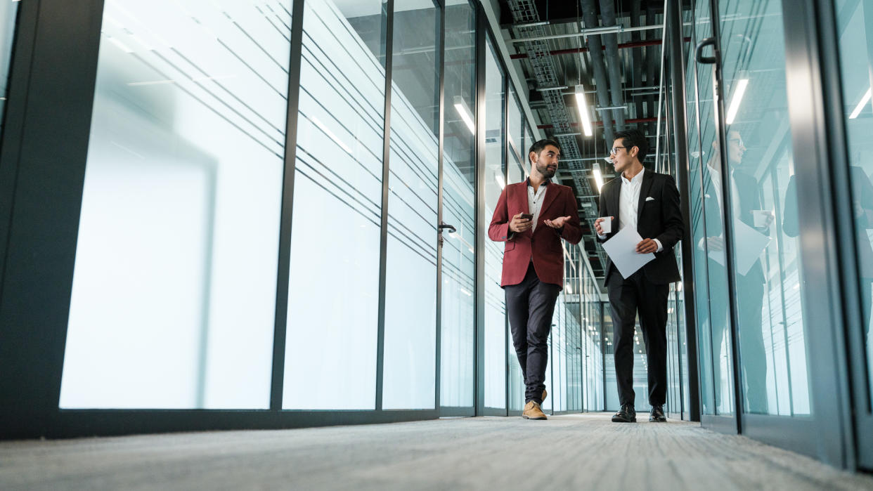 Two corporate men walking in the hallway of a business building and turning to look at each other as one holds a paper and cup and the other gestures with one hand.