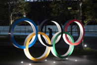 A man is seen through the Olympic rings in front of the New National Stadium in Tokyo, Tuesday, March 24, 2020. IOC President Thomas Bach has agreed "100%" to a proposal of postponing the Tokyo Olympics for about one year until 2021 because of the coronavirus outbreak, Japanese Prime Minister Shinzo Abe said Tuesday. (AP Photo/Jae C. Hong)